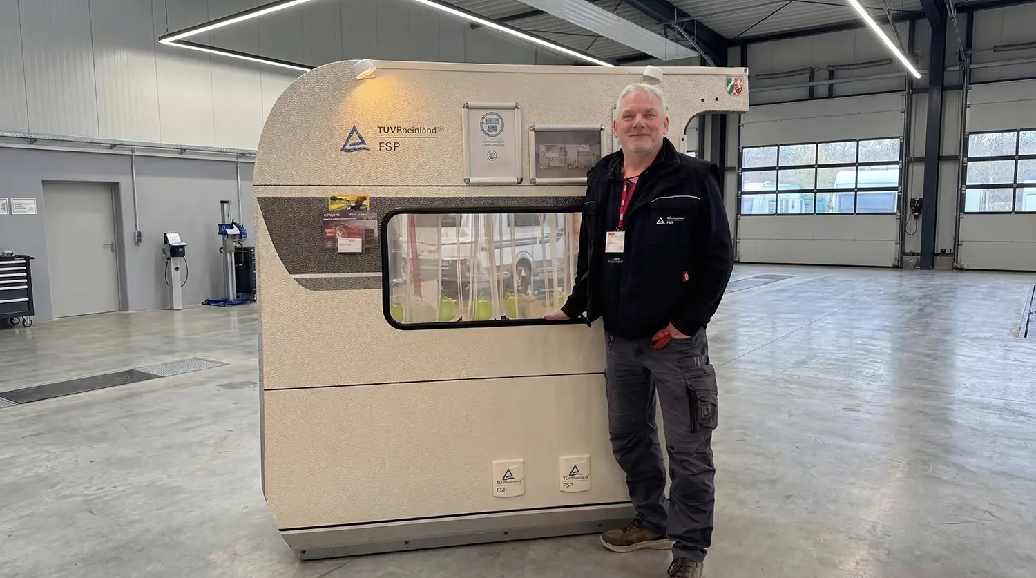 Mann steht in einer modernen Prüfhalle neben einer Caravan Seitenwand mit TÜV Rheinland FSP Kennzeichnung.