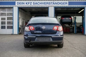 Die Rückansicht eines dunkelblauen VW Passat mit deutlichem Heckschaden steht vor einer Werkstatt die über der Einfahrt das Schild Dein KFZ Sachverständiger trägt während im Hintergrund weitere Fahrzeuge auf Hebebühnen zu sehen sind.