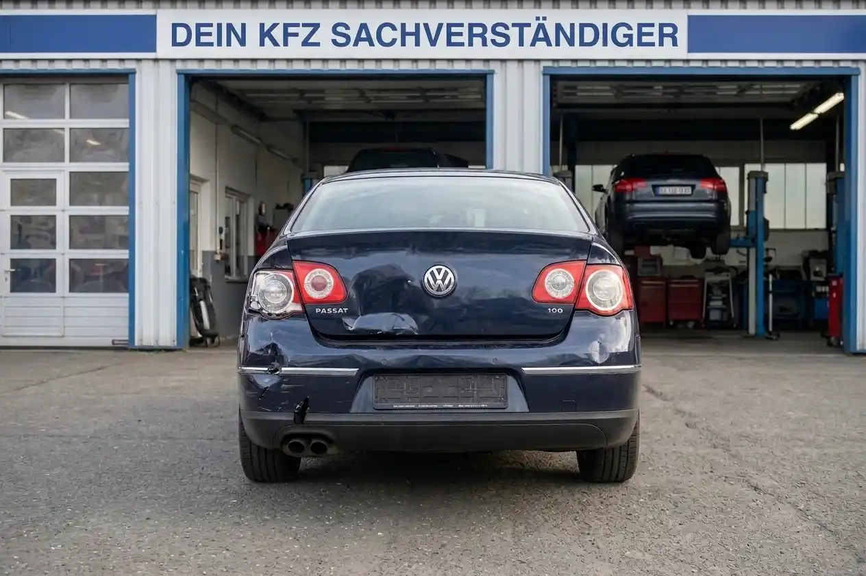 Die Rückansicht eines dunkelblauen VW Passat mit deutlichem Heckschaden steht vor einer Werkstatt die über der Einfahrt das Schild Dein KFZ Sachverständiger trägt während im Hintergrund weitere Fahrzeuge auf Hebebühnen zu sehen sind.
