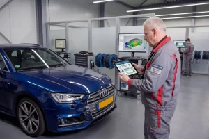 Ein erfahrener KFZ Sachverständiger hält ein Tablet in der Hand und führt eine digitale Schadensbewertung an einem blauen Fahrzeug in einer Werkstatt durch. Im Hintergrund ist ein Monitor mit einer 3D Schadensvisualisierung erkennbar.