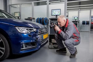 Ein erfahrener Gutachter kniet in einer modern ausgestatteten Werkstatt vor einem blauen Audi und fotografiert mit einer professionellen Kamera einen Frontschaden für das Unfallgutachten