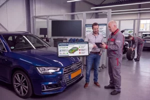 Ein KFZ Sachverständiger bespricht mit einem Kunden in einer modernen Werkstatt eine digitale Schadensanalyse an einem blauen PKW. Im Vordergrund ist ein Monitor mit einer grafischen Auswertung des Fahrzeugzustands zu sehen.