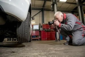 Ein Sachverständiger kniet in einer Werkstatt neben einem angehobenen Fahrzeug und nutzt eine professionelle Kamera um einen KFZ Schaden festhalten zu können.
