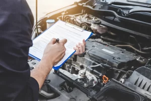 Ein Sachverständiger KFZ dokumentiert die Ergebnisse einer Fahrzeugprüfung auf einem Klemmbrett während er über den offenen Motorraum eines Autos gebeugt ist.