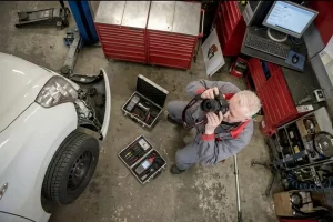 Ein erfahrener Gutachter in Essen fotografiert aus der Vogelperspektive den Frontschaden eines weißen Autos in einer Werkstatt während technische Messgeräte in Koffern neben ihm bereitstehen.