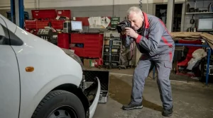 Ein professioneller Sachverständiger steht in einer voll ausgestatteten Werkstatt und fotografiert konzentriert die beschädigte Frontpartie eines weißen Fahrzeugs für eine Kaskoschaden Begutachtung.