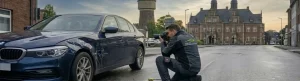 Ein unabhängiger KFZ Sachverständiger von "Dein KFZ Sachverständiger" kniet in einer Straße in Dinslaken und fotografiert einen Unfallwagen, einen dunkelblauen BMW 5er (G30), der einen Frontschaden hat. Im Hintergrund sind Dinslakener Wahrzeichen wie der historische Wasserturm und das Rathaus zu sehen.