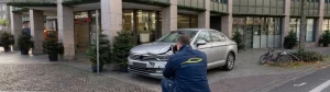 Ein Gutachter von Dein KFZ Sachverständiger kniet vor einem silbernen PKW mit Frontschaden und fotografiert die Beschädigungen. Der Unfallort befindet sich auf einem gepflasterten Vorplatz direkt vor einer Sparkasse Filiale in Mülheim an der Ruhr.